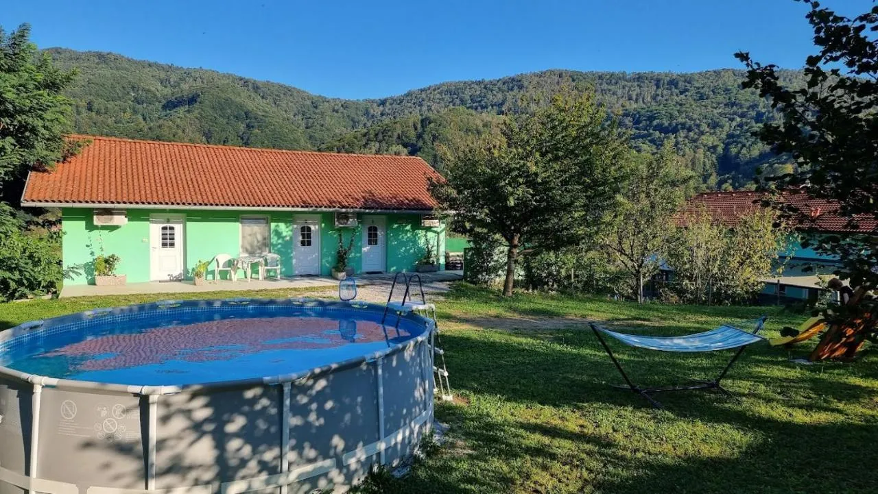 Mintno zelena pritlična hiša z rdečo streho, pred njo travnik z bazenom, zadaj gozdnati hribi