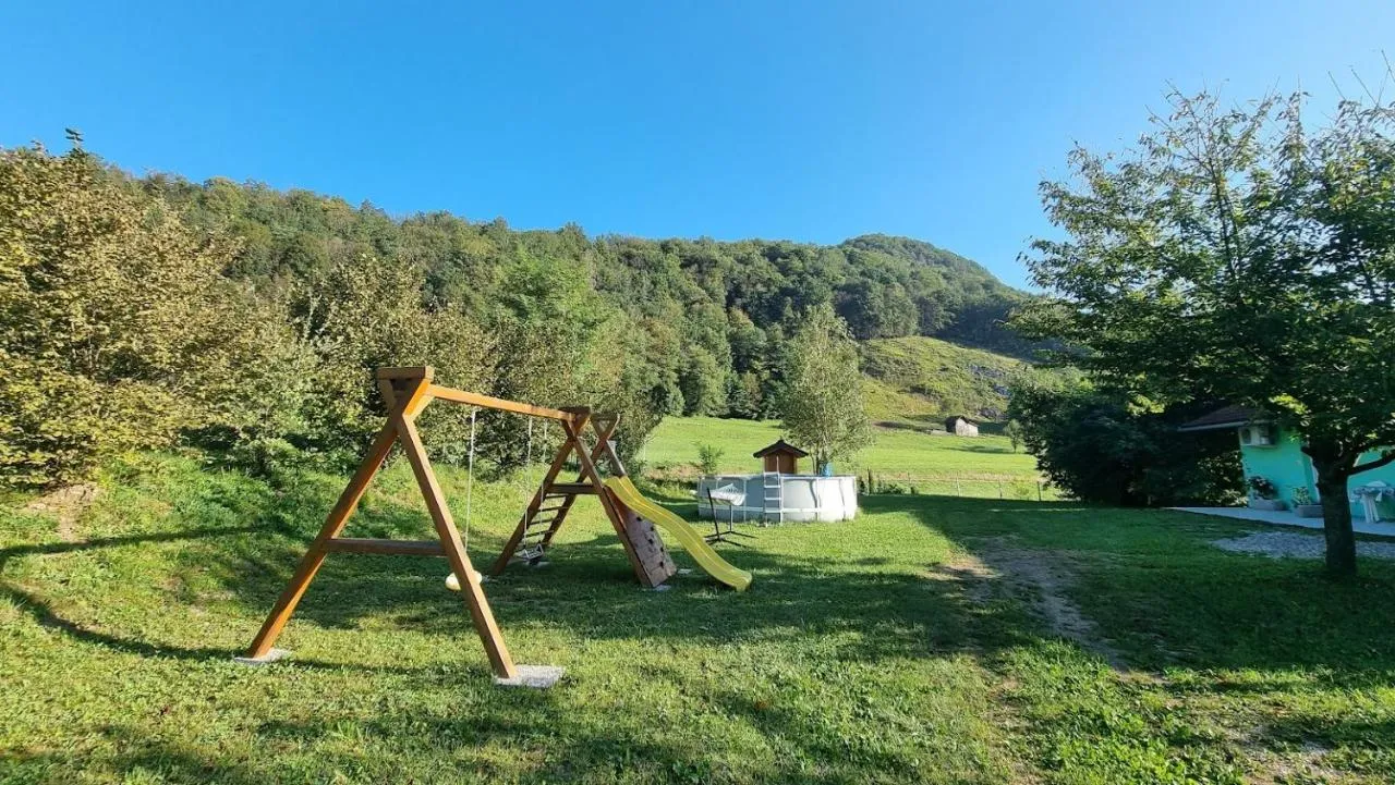Lesena gugalnica s toboganom na travniku, zadaj bazen in gozd